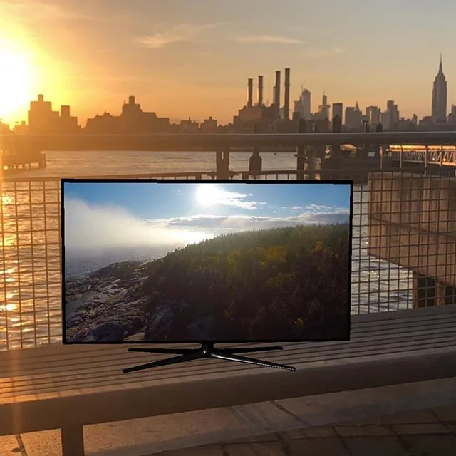 A virtual TV standing on a bench by the water, with the Manhattan skyline behind it at sunset.
